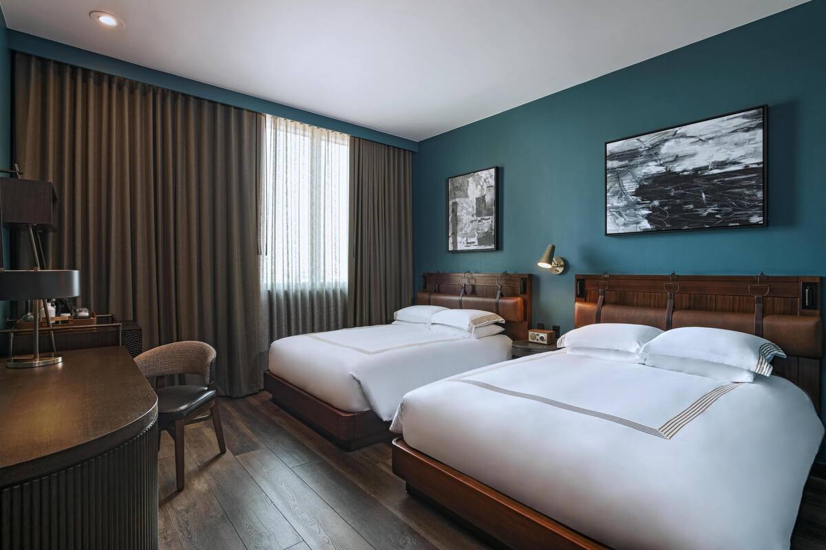 Une chambre d’hôtel avec deux lits, un bureau, une chaise et des œuvres d’art accrochées aux murs.