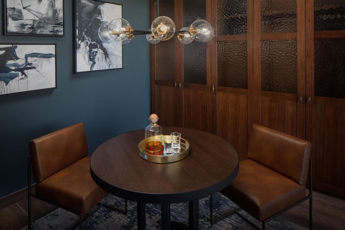 Une table ronde sur laquelle se trouve un plateau avec une bouteille, des verres et une bougie. Deux fauteuils en cuir sont placés autour de la table.
