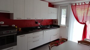 A modern kitchen with white cabinets, a black countertop, and a red accent wall. There is a dining table with chairs and a window with red curtains.