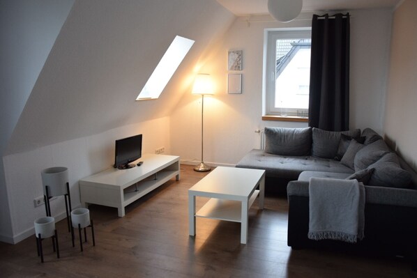Ein modernes Wohnzimmer im Loft-Stil mit schrägverdeckter Decke, einem Fernseher auf einer weißen Konsole, einem grauen Sofa, einem weißen Couchtisch und einer Stehlampe.