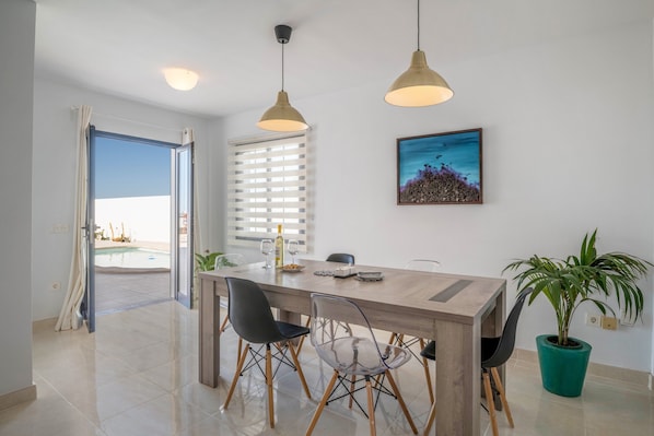 Un coin repas avec une table en bois, des chaises noires et transparentes, des luminaires suspendus, une plante en pot et un tableau encadré au mur.