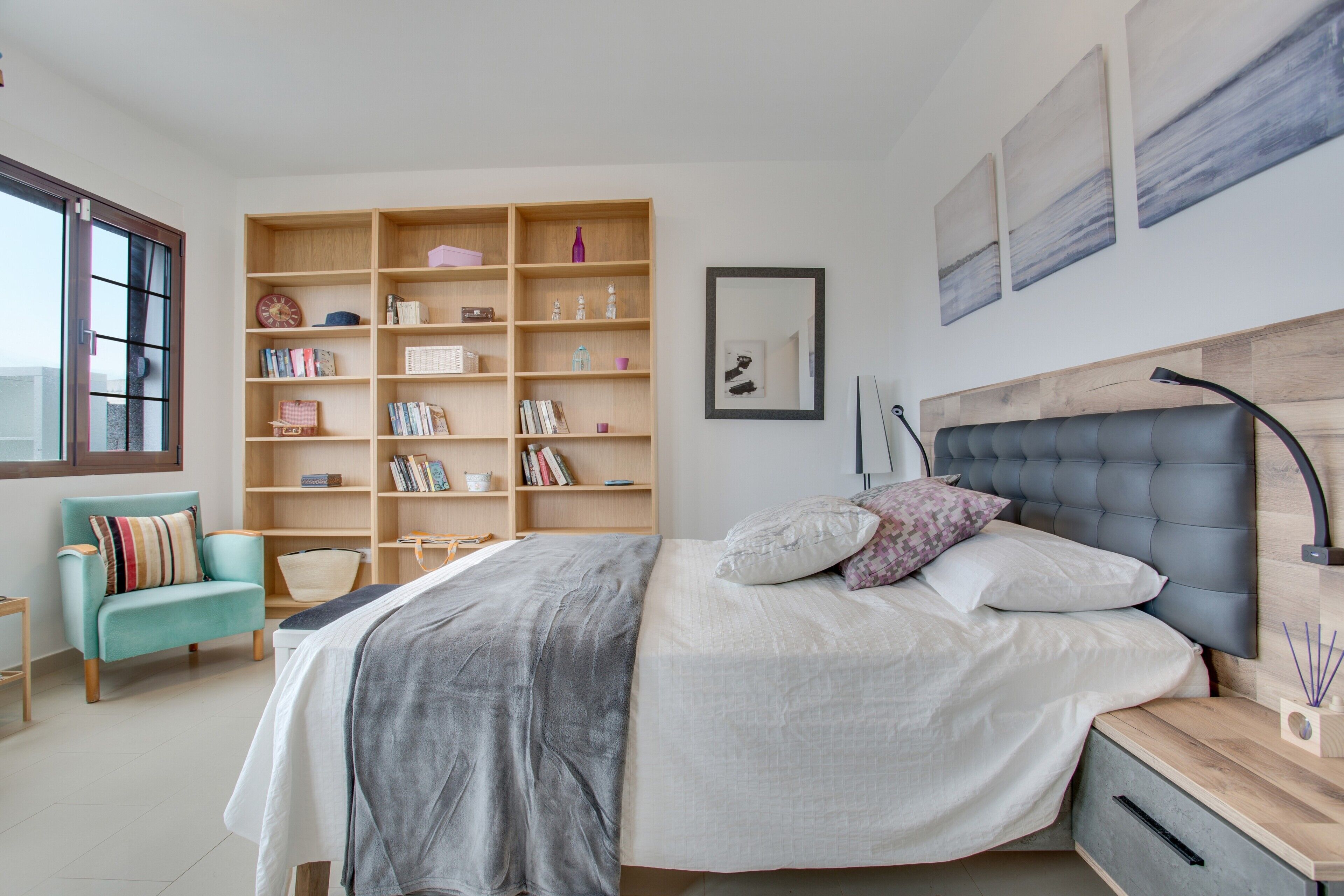 A bedroom with a bed, bedside table, bookshelf, and a window with blinds.