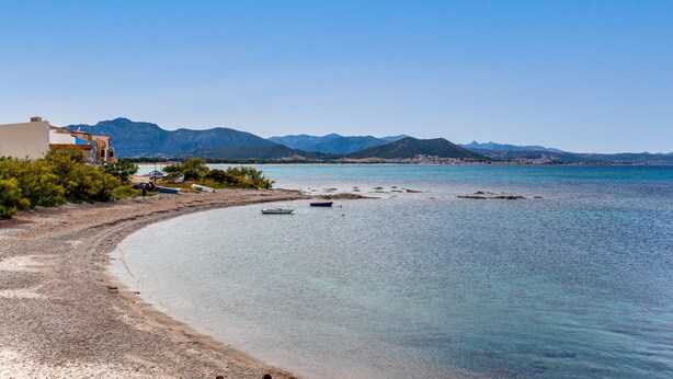A coastal landscape with a beach, clear water, and mountains in the background.