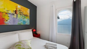Ein Hotelzimmer mit einem Bett, einem Nachttisch mit Lampe, einem Fenster mit Vorhängen und einem farbenfrohen abstrakten Gemälde an der Wand.