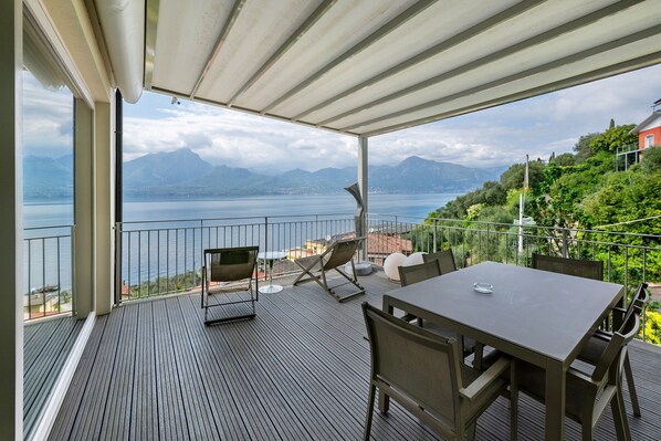 A covered outdoor area with a table and chairs, overlooking a body of water and mountains.