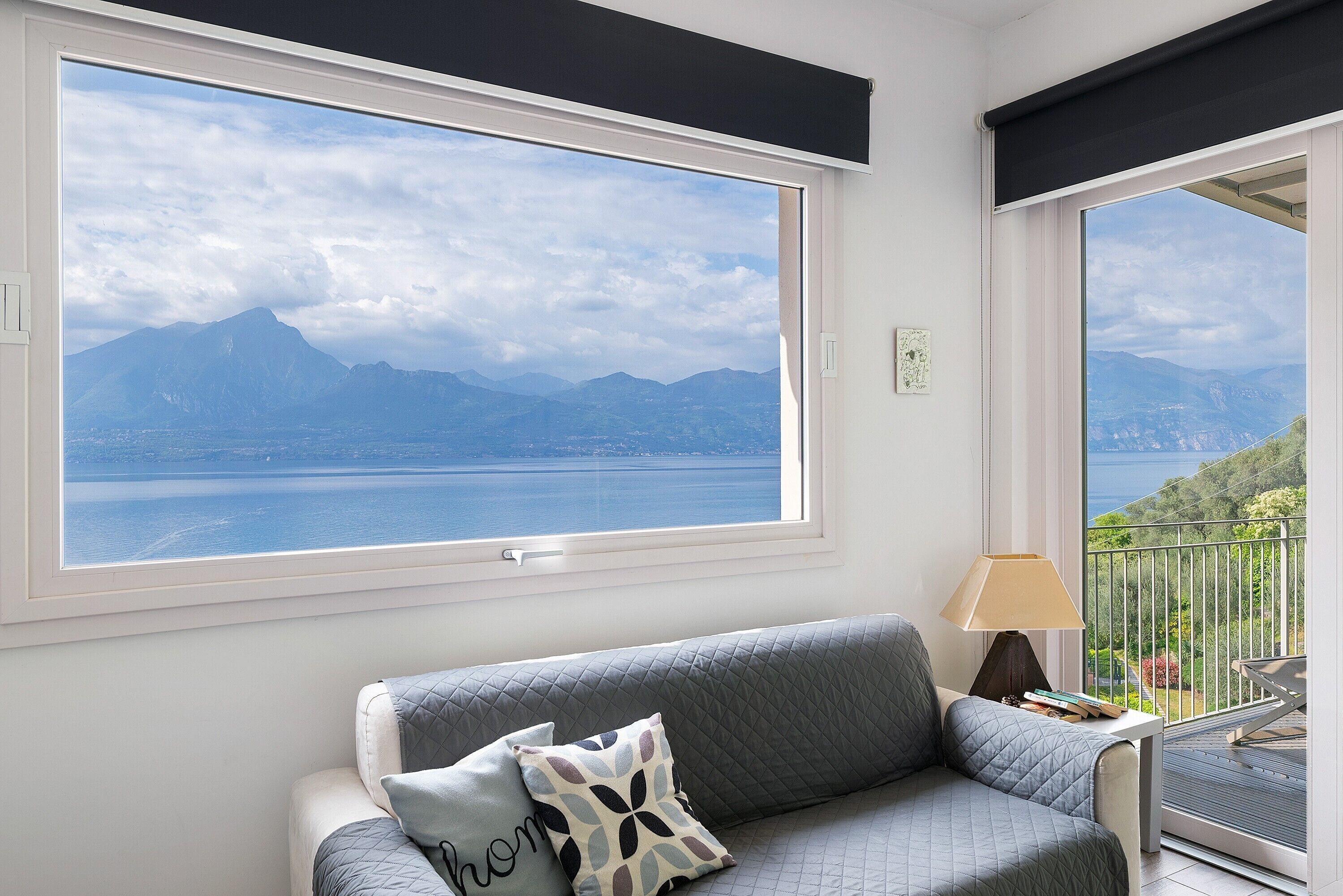 Ein Zimmer mit großem Fenster und Blick auf die Berge, ein graues Sofa mit gemusterten Kissen sowie ein kleiner Tisch mit einer Lampe.