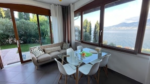 Un salon moderne avec une table à manger en verre, des chaises blanches, un canapé beige agrémenté de coussins à motifs et de grandes fenêtres offrant une vue sur un lac et des montagnes.