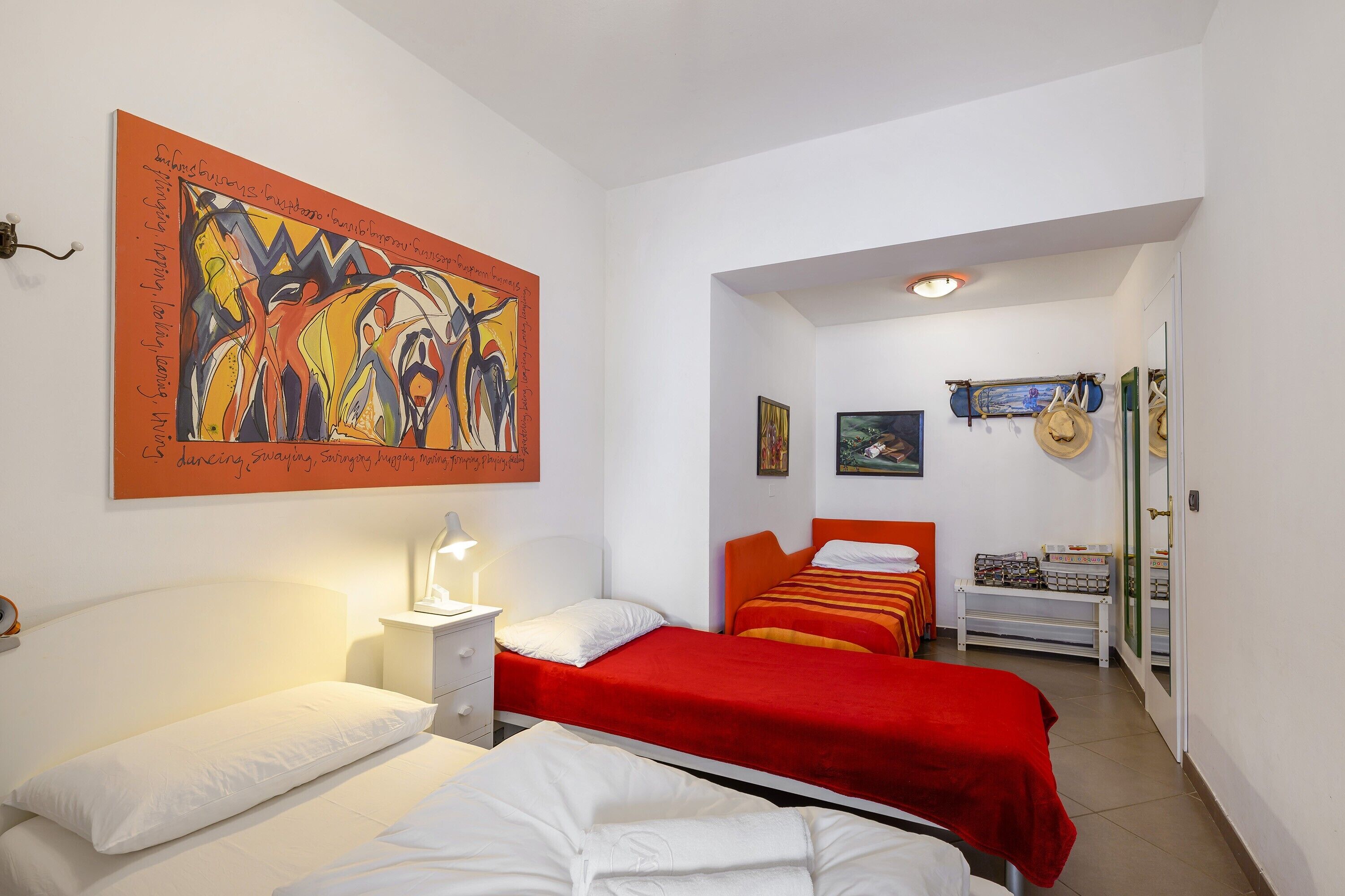Une chambre d’hôtel avec deux lits, un tableau au mur, une table de chevet et un banc sur lequel sont posés des chapeaux.