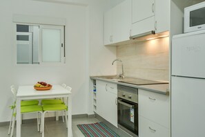 A kitchen with white cabinets, a stainless steel sink, a microwave, and a stove. There is a dining table with green chairs and a bowl of fruit. - Cozy Apartment "1B SRV" Near the Sea with Wi-Fi and Fans (VECINDARIO)
