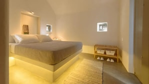 A bedroom with a bed, bedside tables, a nightstand, a small wooden bench, and a wall-mounted shelf with baskets. - Salty villa with private Pool & Sea view (Santorini)