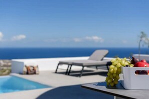 Un área junto a la piscina con una tumbona, una mesa con frutero y vista al océano.