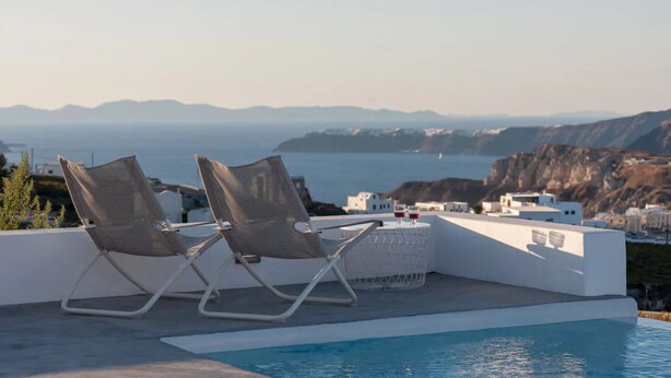 A poolside area with two lounge chairs and a table, overlooking a coastal landscape with buildings and cliffs. - Sandy villa with private pool & Sea view (Santorini)