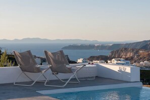 A poolside area with two lounge chairs and a table, overlooking a coastal landscape with buildings and cliffs. - Sandy villa with private pool & Sea view (Santorini)