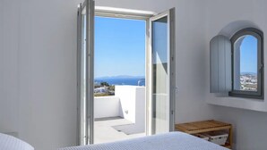A bedroom with a bed, a wooden bench, a window, and a balcony with a view of the sea and buildings. - Sandy villa with private pool & Sea view (Santorini)
