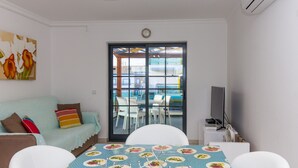 A room with a sofa, a dining table with chairs, a TV, and a large window with a view of an outdoor pool area.
