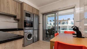 A modern kitchen with a built-in oven, microwave, and washing machine, a dining area with a wooden table and red chairs, and a sliding glass door leading to a balcony with a view of buildings.