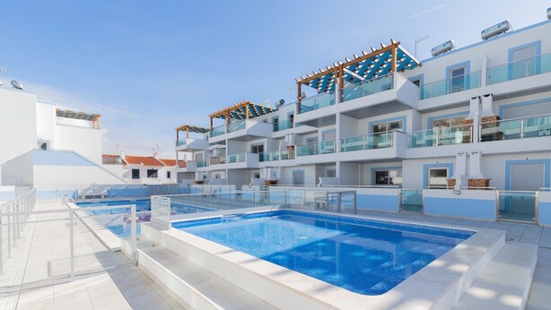 A modern hotel with a large outdoor swimming pool, multiple balconies, and a clear blue sky.
