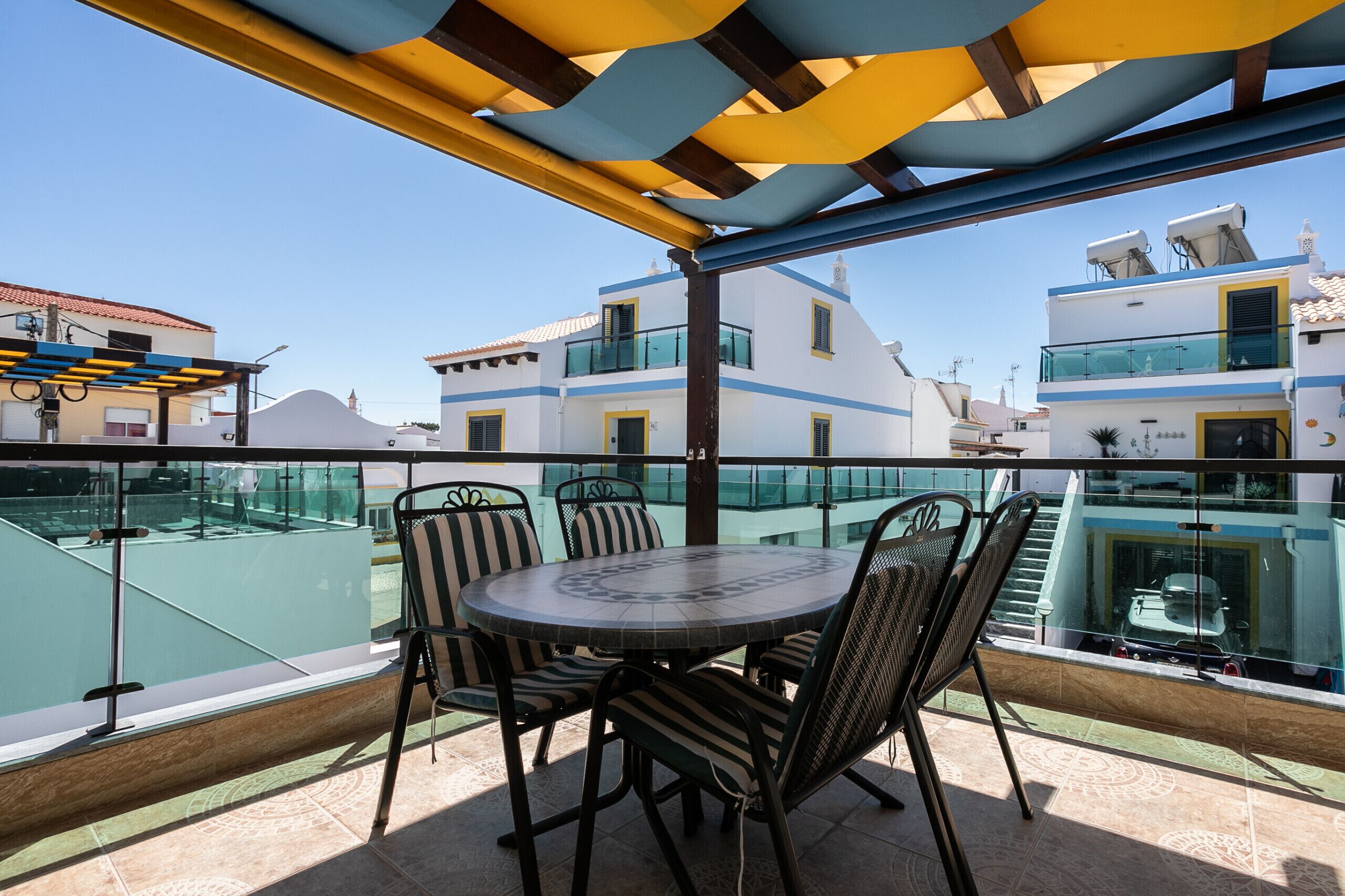 A terrace with a round table and chairs, overlooking a pool and buildings.