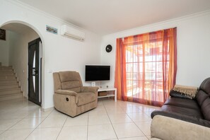 A living room with a beige sofa, a brown leather sofa, a recliner, a flat-screen TV on a white stand, a window with orange curtains, and a staircase leading to another room.