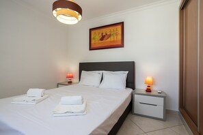 A neatly made bed with white linens and towels, two bedside tables with lamps, a headboard, and a framed painting on the wall.