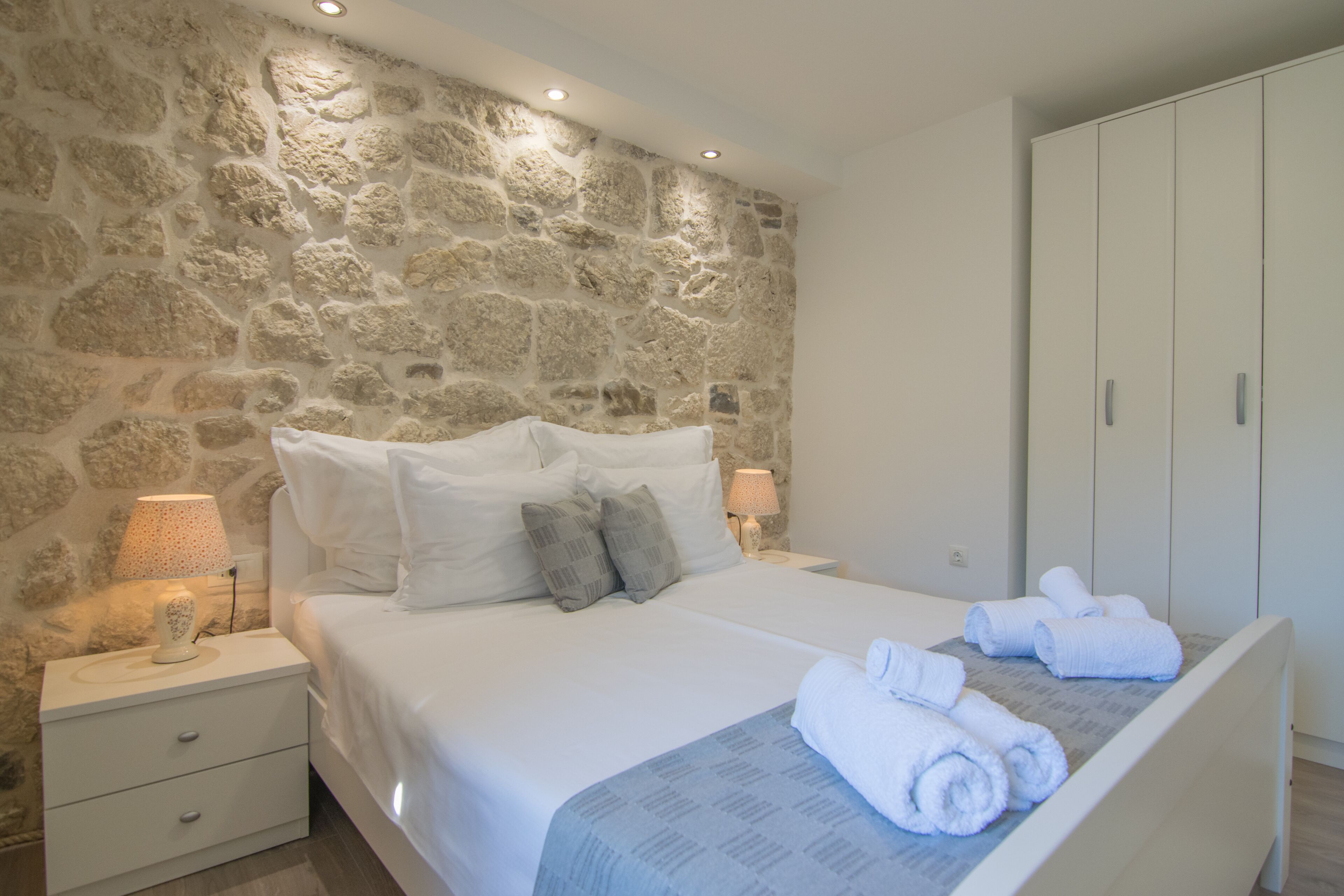 A bedroom with a stone wall, a bed with white bedding, two bedside tables with lamps, and a wardrobe.