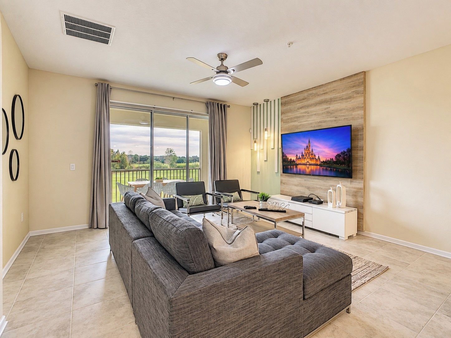 A modern living room with a large sofa, a flat-screen TV, a ceiling fan, and a view of the outdoors.