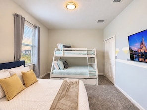 A hotel room with a bunk bed, a single bed, a large window with curtains, a flat-screen TV, and a painting of a castle.