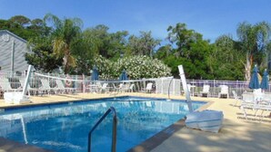Una piscina con una escalera metálica, rodeada de sillas reclinables y sombrillas blancas, con árboles y un edificio al fondo.