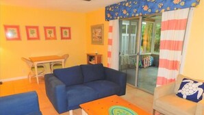 Una sala de estar con un sofá azul, una mesita de centro naranja y un sillón gris. Hay cuadros enmarcados en la pared y una puerta de cristal que da al exterior.