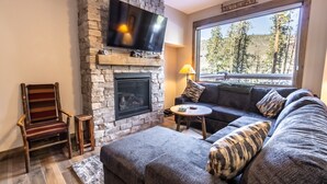 A cozy living room with a stone fireplace, a flat-screen TV mounted above it, a large window with a view of trees, a comfortable sofa, and a wooden chair. - New Complex Close to Shuttle for Easy Access to Resort w/ Private Hot Tub! (Keystone)