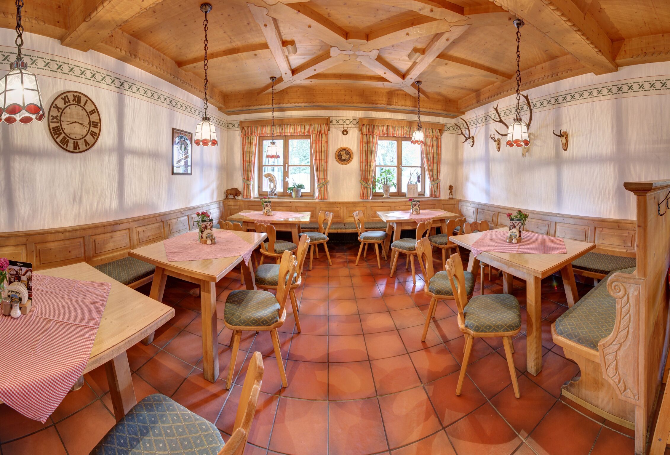 Uma área de jantar rústica com mesas e cadeiras de madeira, um relógio grande na parede e uma janela com cortinas.