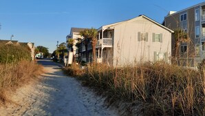 A sandy path leading to a building with a balcony, surrounded by tall grasses and a clear blue sky.