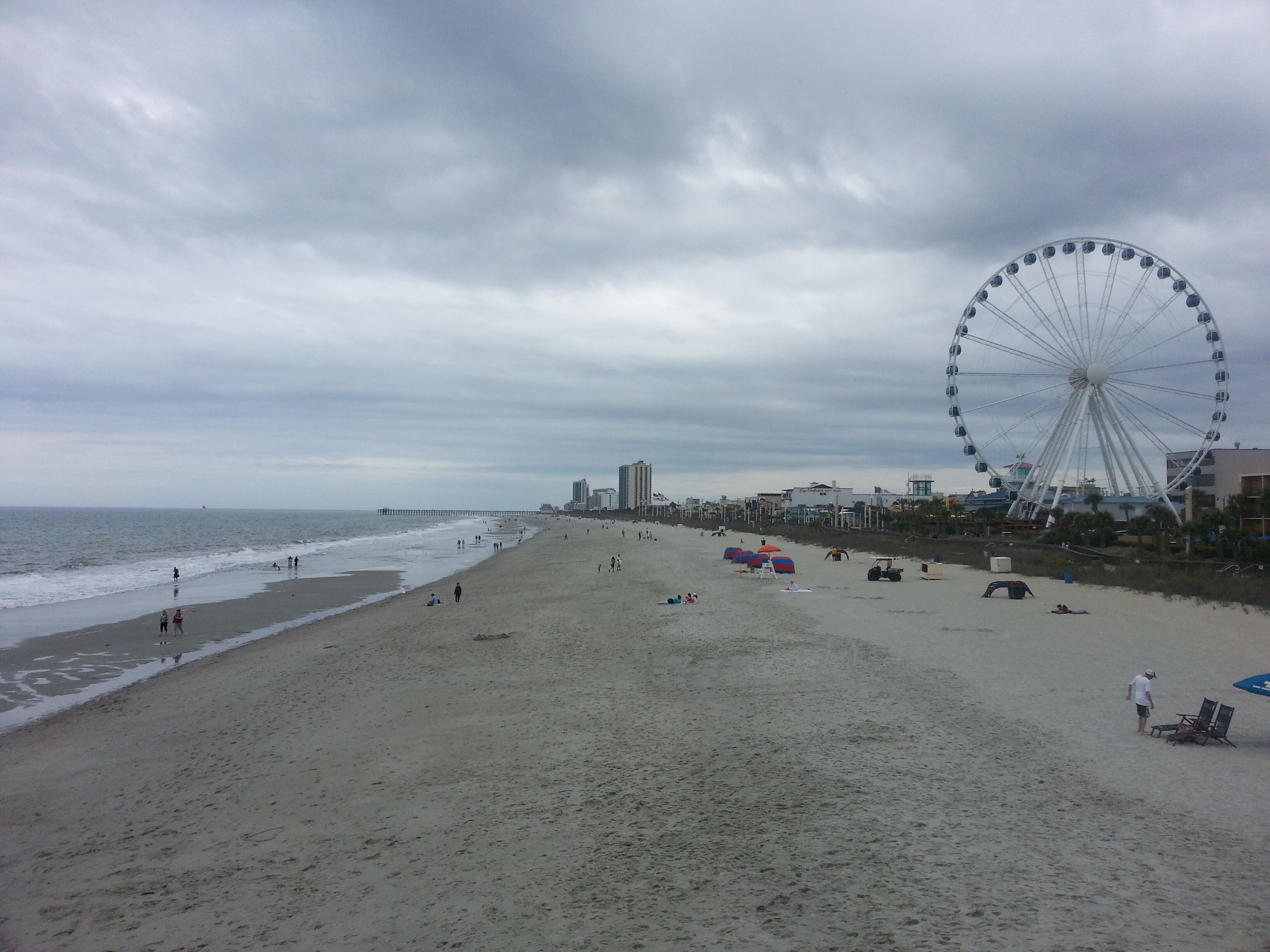 A beach with a large Ferris wheel in the background, a pier extending into the ocean, and a few beachgoers.