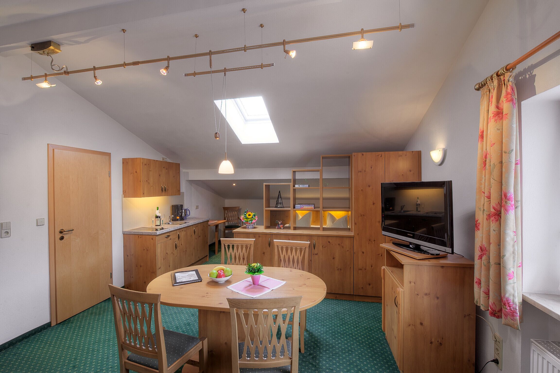 A compact kitchen with wooden cabinets, a dining table with chairs, a TV, and a small window with a curtain.