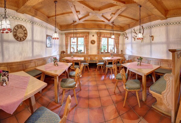 Un coin repas rustique avec des tables et des chaises en bois, une grande horloge au mur et une fenêtre avec des rideaux.