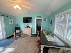 A room with teal walls, a ceiling fan, a flat-screen TV, a dining table with chairs, a window with blinds, and a door leading to another room.