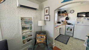 A compact living space with a kitchenette, a sofa with a patterned cushion, and a wall-mounted air conditioner.