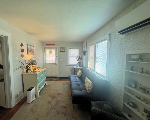 A living room with a sofa, a small table with a lamp, and a shelf with decorative items.