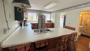 A kitchen with a central island, wooden bar stools, a refrigerator, and a microwave. - Miss Agnes - Water Front 3BR home PET FRIENDLY, NO LINENS PROVIDED (Chincoteague)