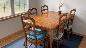 A dining room with a wooden table and chairs, a painting on the wall, and a window with a view of trees.