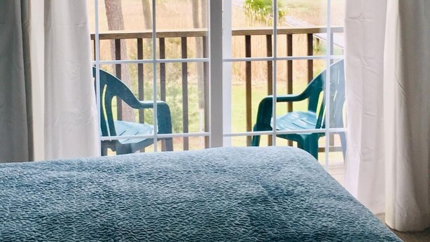 A room with a bed, a window with white curtains, and a view of outdoor chairs and trees.