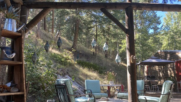Un área de estar al aire libre de estilo rústico, con sillas y mesas de madera, rodeada por una pérgola de madera y faroles colgantes.