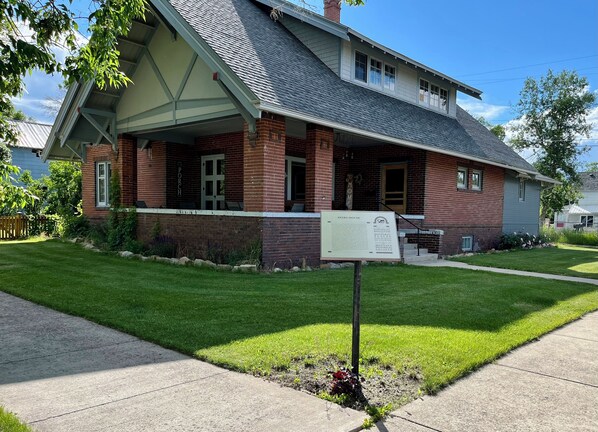 A two-story brick house with a porch, a well-maintained lawn, and a signboard in front.