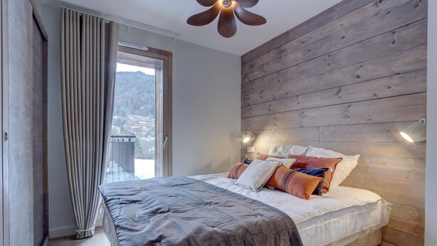Un dormitorio con cabecera de madera, una cama grande con muchas almohadas, una ventana con cortinas y vistas a un paisaje montañoso.