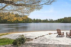 A lakeside setting with a sandy beach, two Adirondack chairs, and a forested backdrop.