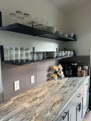 A kitchen with a marble countertop, a black coffee machine, and various glassware and dishes.