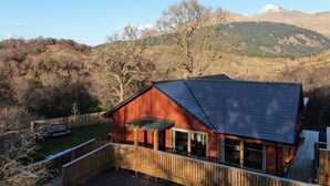 A wooden cabin with a deck, large windows, and a mountain backdrop. - Hot tub lodge with stunning view (Taynuilt)