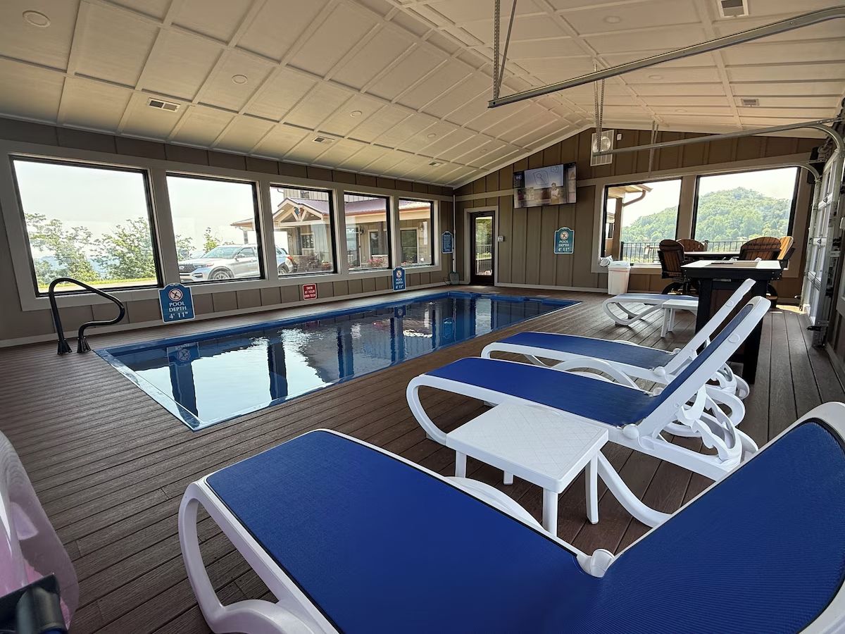 A pool area with lounge chairs and a table, large windows, and a view of the outside.