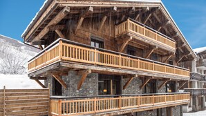 A multi-story wooden chalet with balconies, stone foundation, and a snow-covered landscape.