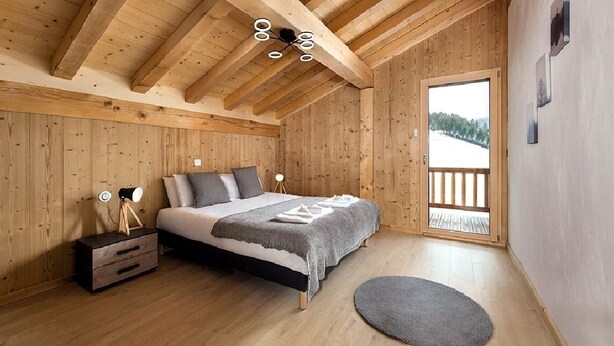 A wooden-paneled bedroom with a bed, bedside tables, and a view of a snowy landscape through a large window.